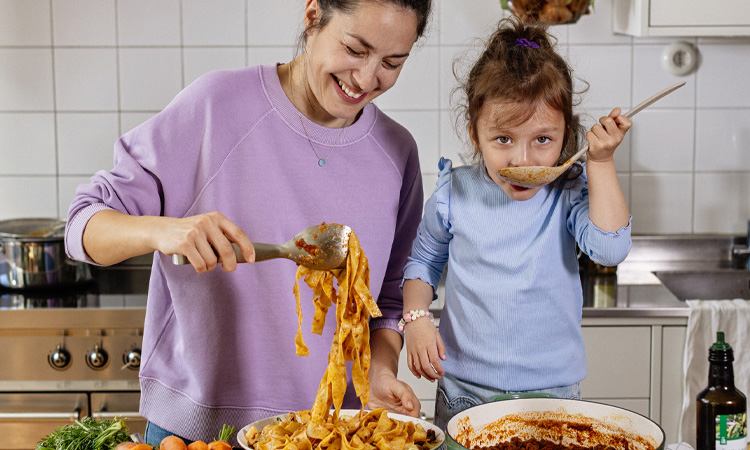en kvinna och ett barn står i ett kök och håller i pasta slevar och lyfter upp spagetti ur en skål